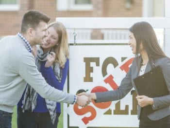 A Caucasian man and woman are buying a house together. They are standing outside the house with a sold sign and their real estate agent. The man and agent are shaking hands.